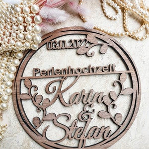 May include: A round wooden sign with the date "03.10.2022" and the text "Perlenhochzeit Karin & Stefan" carved into it. The sign is decorated with a leafy design.