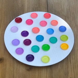 May include: A white circular board with 25 small, colorful, round paint pots arranged in a circular pattern. The paint pots are arranged in a rainbow color order, starting with red and ending with yellow.