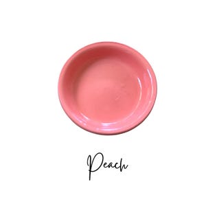 May include: A small, round, peach-colored ceramic bowl. The bowl has a slightly raised rim and a smooth interior. The word "Peach" is written in cursive script below the bowl.