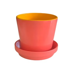 May include: A coral-colored ceramic flower pot with a matching saucer. The interior of the pot is painted a bright yellow. The pot has a slightly tapered shape, wider at the top and narrower at the base.