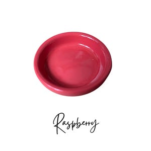 May include: A small, round, red ceramic dish. The dish is smooth and has a glossy finish.