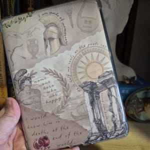 May include: A clear tablet case featuring a beige background with Greek-themed illustrations. The design includes a helmet, a statue, a sunburst, and architectural elements, along with handwritten text. The case is designed to protect the tablet.