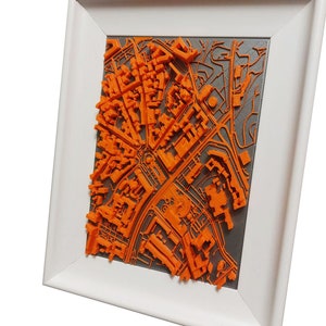 May include: A 3D printed cityscape model in a white frame. The model is orange and depicts a city with buildings, roads, and parks. The frame is white with a grey mount.