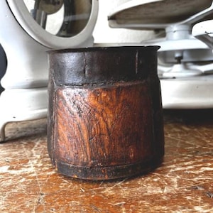 May include: A cylindrical wooden container with a dark, weathered appearance. The top and bottom rims are a darker, almost black color, contrasting with the lighter, textured wood grain of the body. The container sits on a wooden surface.