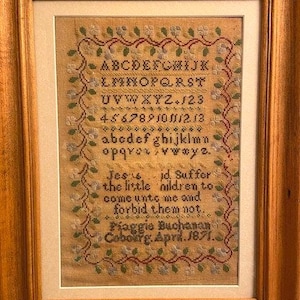 May include: A framed sampler with a cream-colored background and a colorful floral border. The sampler features the alphabet, numbers, and a verse from the Bible: "Jesus, I'd suffer the little children to come unto me and forbid them not." The sampler is signed "Piaggie Buchanan, Cobourg, April 1871."