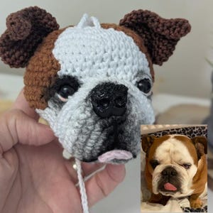 May include: A crocheted bulldog head ornament in white, brown, and grey, with black eyes and nose. The handmade ornament features a pink tongue and is a replica of a real bulldog.