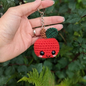 May include: A red crocheted apple keychain with a smiling face, black eyes, and pink cheeks. The apple has a brown stem and a green leaf. The keychain is held up against a background of green foliage.