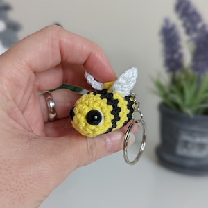 May include: A crocheted bee keyring. The bee has a yellow body with black stripes, large black eyes, and white wings. The keyring is attached to a silver metal ring.