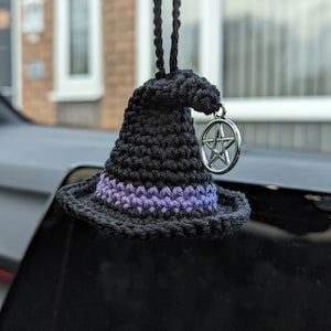 May include: A black crochet witch hat car charm with a purple band and a silver pentacle charm. The hat is hanging from a black cord. The hat is a decorative item.