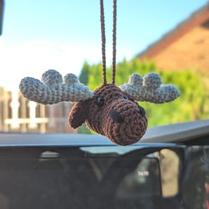 May include: A crocheted moose ornament featuring a dark brown head with black plastic eyes and a black stitched nose. The moose has large, textured white antlers and is suspended by a thin brown crocheted cord for hanging as a decoration.
