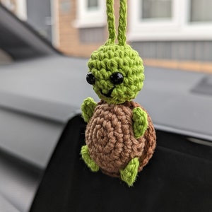 May include: A crocheted turtle car charm, featuring a green head and limbs, a brown shell, and black eyes. The charm is suspended by a green cord. This handmade item is designed to hang from a car's rearview mirror.