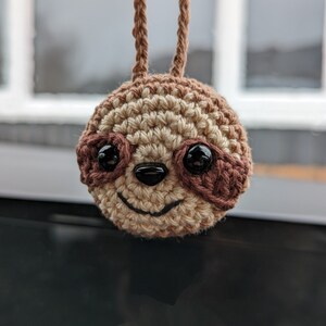 May include: A crocheted sloth head with a smiling face and dark, glossy eyes. The sloth is made of tan and brown yarn, with a small, black nose and a stitched mouth. Two brown yarn loops extend from the top of the head.