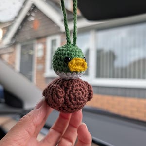May include: A small, handmade crochet mallard duck ornament featuring a green head, a yellow beak, a white neck ring, and a brown body. The duck has black bead eyes and is attached to a green yarn loop for hanging as a decorative accessory.