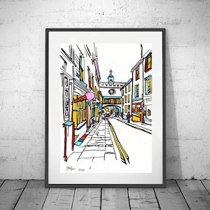 Puede incluir: Una impresión artística enmarcada de una escena callejera. La obra de arte representa una vista colorida y estilizada de una calle europea con edificios, tiendas y una torre del reloj. La impresión está en un marco negro y se apoya en una pared de ladrillos blancos.
