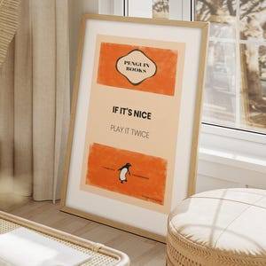Op de afbeelding: Een ingelijste poster met een lichtbeige achtergrond en oranje accenten. De poster bevat de tekst "Penguin Books" en "If it's nice, play it twice". Een pinguïn illustratie is ook inbegrepen. De lijst heeft een lichte houtkleur.
