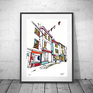 Puede incluir: Una impresión artística enmarcada de una escena callejera con edificios y escaparates coloridos. La obra de arte presenta contornos negros audaces y colores vibrantes, incluyendo rojo, naranja y azul. La impresión se muestra en una pared blanca.