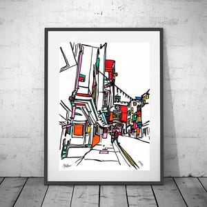 Puede incluir: Una impresión artística enmarcada de una escena callejera con edificios en un estilo abstracto colorido. La obra presenta contornos negros audaces con toques de rojo, naranja, rosa y turquesa. La impresión se exhibe en una pared blanca y un suelo de madera.