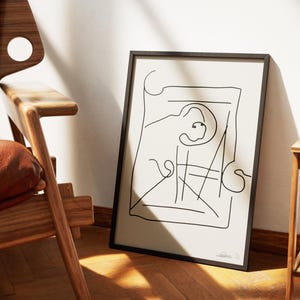 May include: Abstract line art print in a black frame, displaying a minimalist design on a cream backdrop. The artwork is propped against a white wall, beside a wooden chair with a brown leather seat. The room has a warm atmosphere.