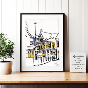 Puede incluir: Dibujo en blanco y negro de una escena de calle con acentos de color. El dibujo representa una fila de edificios con ventanas y puertas. Los edificios son de estilo europeo con un techo puntiagudo. El dibujo está enmarcado en un marco negro. El tamaño de la impresión enmarcada es de 16,5x23,4 pulgadas o 42x59 cm.