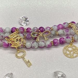 May include: Three-strand white and purple beaded Apple Watch band with gold charms. The charms include a key, a bow, and a circle with the word "LOVE" inside.
