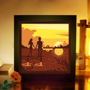 May include: A framed, illuminated art piece showing a couple silhouetted on a pier, holding hands, with a sunset over a lake. A boat is on the water, and the sky is orange and yellow. A rose is in the foreground.