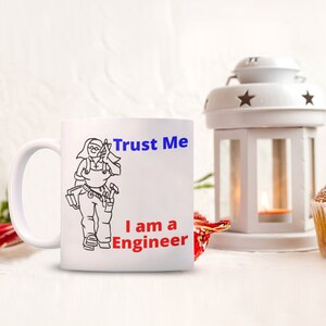 May include: White ceramic mug with a black line drawing of a woman wearing a tool belt and holding a wrench. The text "Trust Me I am a Engineer" is printed in blue and red on the mug.
