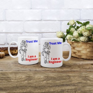 May include: Two white ceramic mugs with black outlines of a person holding tools. The text on the mugs reads "Trust Me I am a Engineer" in blue and red.