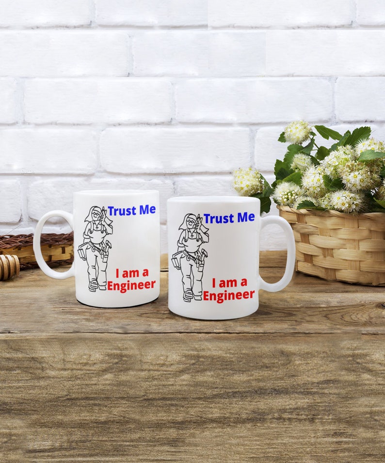 May include: Two white ceramic mugs with black outlines of a person holding tools. The text on the mugs reads "Trust Me I am a Engineer" in blue and red.