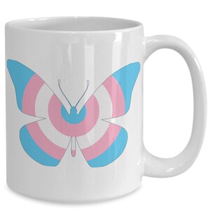 Puede incluir: Taza de cerámica blanca con un diseño de mariposa azul y rosa. La mariposa tiene un patrón de bandera del orgullo transgénero.