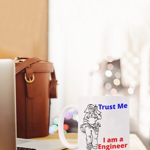 May include: White ceramic mug with a black line drawing of a person wearing a tool belt and holding a wrench. The text "Trust Me I am a Engineer" is printed on the mug in blue and red.
