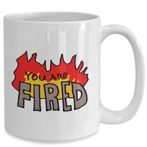 May include: White ceramic coffee mug with a red, yellow, and gray graphic that says "You are Fired" in a cartoon style.