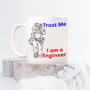 May include: White ceramic mug with a black line drawing of a person wearing overalls and holding tools. The text "Trust Me I am a Engineer" is printed in blue and red on the mug.