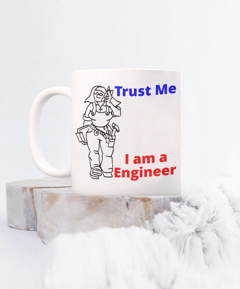 May include: White ceramic mug with a black line drawing of a person wearing overalls and holding tools. The text "Trust Me I am a Engineer" is printed in blue and red on the mug.
