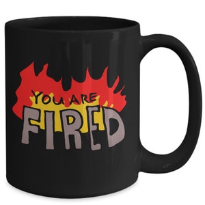 May include: Black ceramic coffee mug with a white and gray graphic of flames and the text "You are Fired" in yellow and red.