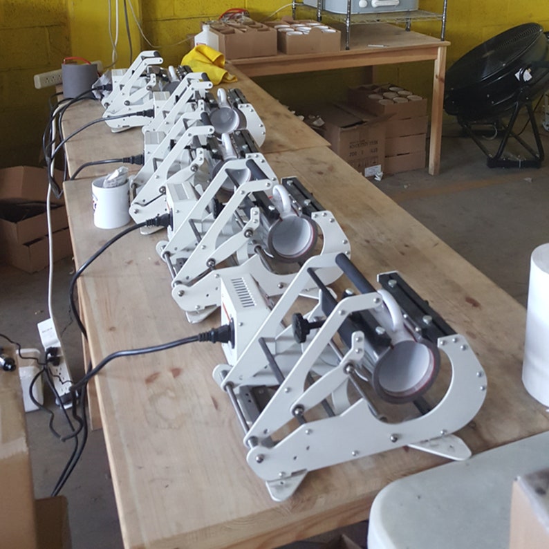 May include: A row of six white mug presses with black handles and cords plugged into a power strip. Each press has a white ceramic mug inside. The presses are on a wooden table.