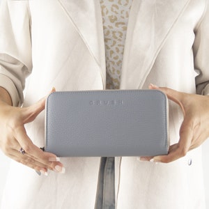 Könnte beinhalten: Hellblaues Lederportemonnaie mit dem Wort "CRUSH" auf der Vorderseite. Das Portemonnaie hat einen Reißverschluss und wird von einer Person gehalten. Der Hintergrund umfasst eine weiße Jacke und ein gemustertes Hemd.