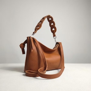 May include: A tan handbag with a pebbled texture, featuring a top zip closure, a knotted side detail, and a decorative chain-link style handle. The bag includes a long, thin, adjustable shoulder strap attached with silver-toned metal hardware.