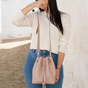 May include: A light pink leather bucket bag with a drawstring closure and a long strap. The bag is worn over the shoulder of a person wearing a white sweater and blue jeans.