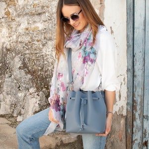 May include: A light blue bucket bag with a drawstring closure and a long strap. The bag is worn by a person wearing a white shirt with a floral scarf and blue jeans.