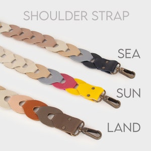 Può includere: Cinturini a tracolla in pelle intrecciata in vari colori: beige, grigio, rosa, giallo e marrone. Ogni cinturino ha un moschettone in metallo. Il testo "SHOULDER STRAP" è in alto, con "SEA", "SUN" e "LAND" sotto.