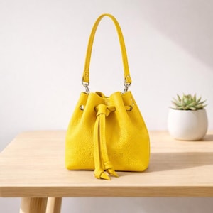 Può includere: Una borsa a secchiello giallo brillante con manico e chiusura a coulisse abbinati. La borsa ha una superficie strutturata e finiture color argento. La parola "CUSH" è in rilievo sul davanti. Una piccola pianta grassa in un vaso bianco è sullo sfondo.