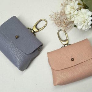 Puede incluir: Dos pequeñas bolsas de cuero texturizado en azul polvoriento y rosa rubor. Cada bolsa tiene un cierre de color latón y un cierre a presión. Las bolsas se muestran sobre una superficie blanca, con flores blancas en el fondo.