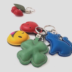 May include: A collection of leather keychains in various shapes and colors. Includes a red cherry, a red ladybug, a yellow smiley face, a green four-leaf clover, and a blue cat. Each keychain has a silver metal ring.