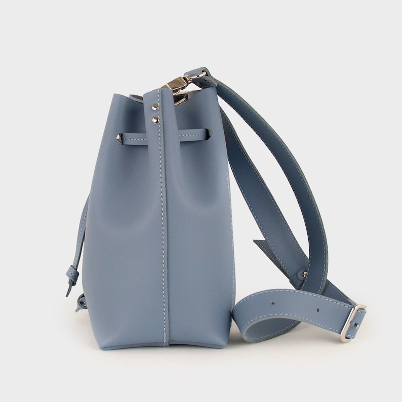 May include: A light blue leather bucket bag with a long adjustable strap. The bag has a drawstring closure and a silver buckle.