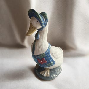 May include: A ceramic duck figurine, wearing a blue and green plaid bonnet, vest, and scarf. The duck has a yellow beak and stands on a grey base. A red flower adorns the vest.