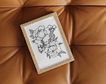 Op de afbeelding: Een ingelijste zwart-wit bloemenkunstprint met pioenrozen. Het kunstwerk is geplaatst in een lichtbruine, getextureerde lijst. De print ligt op een bruin lederen oppervlak.