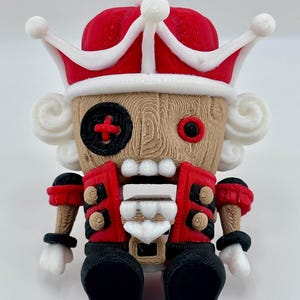 Puede incluir: Una figura de cascanueces roja y blanca con una corona, un parche en el ojo negro y un uniforme rojo y negro. La figura está sentada con las piernas cruzadas.