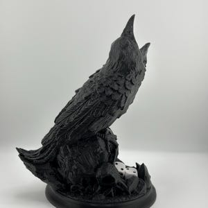 May include: A black 3D printed sculpture of two crows perched on a skull with dice at their feet.