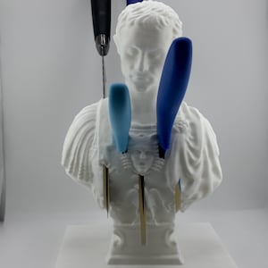 以下が含まれることがあります： 胸に4本のナイフが刺さった男性の白いバスト彫刻。ナイフは青と水色のハンドルです。