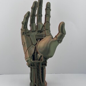 May include: A bronze-colored, robotic hand with intricate mechanical details. The fingers and palm are articulated, showcasing a blend of organic and technological design. The hand is posed in an open gesture, highlighting its complex construction.